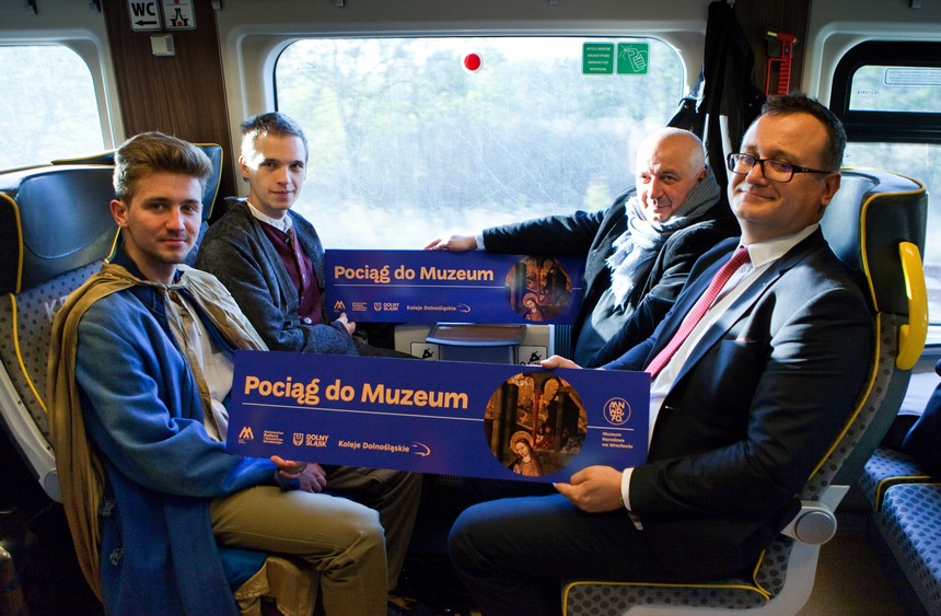 Foto: Dyrektor Muzeum, Prezes Zarządu KD i aktorzy siedzą w pociągu i trzymają tablicę z napisem Pociąg do Muzeum