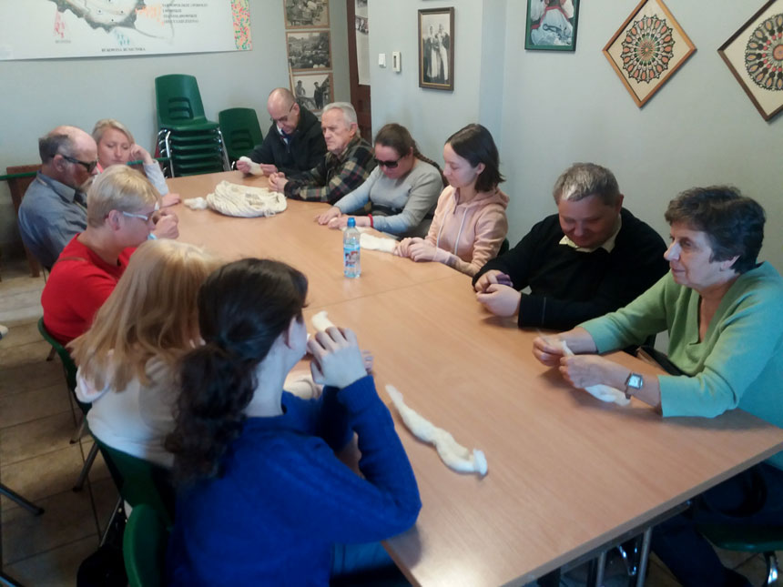 foto: uczestnicy zajęć siedzą wokół dużego stolu i z wełny rolują wałki, z których będzie tworzony filc – inne ujęcie, przed uczestniczą w granatowym sweterku leży pasek białej wełny