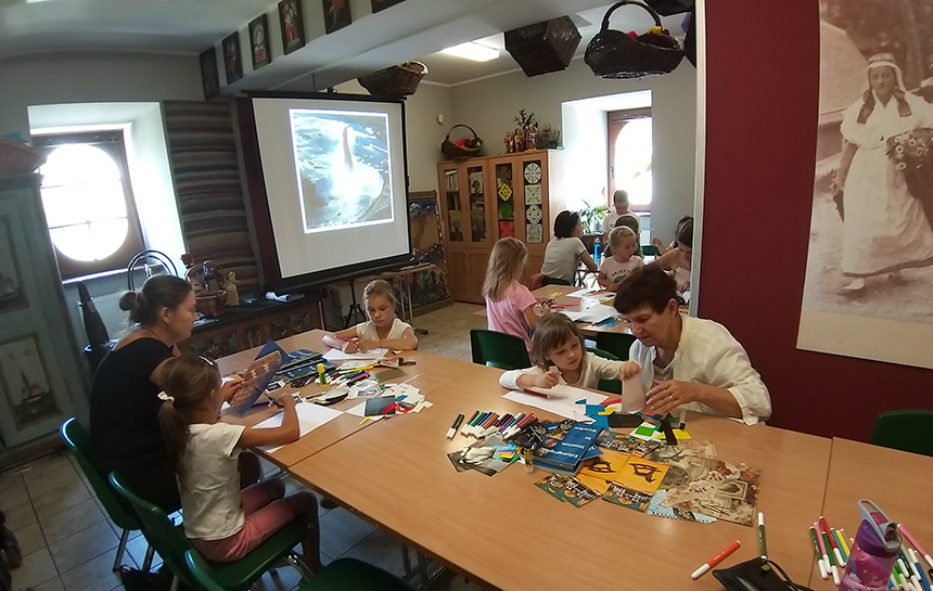 zajęcia w salce edukacyjnej, uczestnicy siedzą przy stołach i przeglądają materiały do kolażu – skrawki kolorowych gazet i folderów
