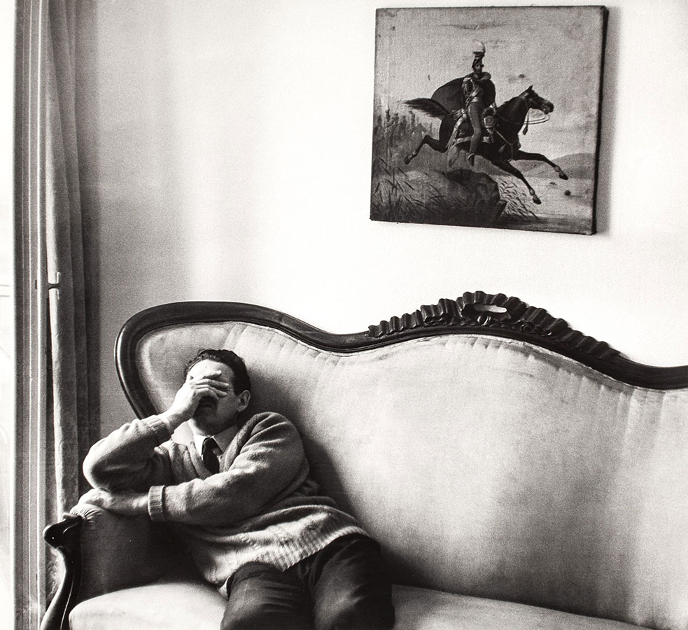 czarno-biała fotografia przedstawia siedzącego na kanapie reżysera Andrzeja Wajdę, który zmęczony przesłania dłonią oczy, na ścianie w tle obraz z ułanem