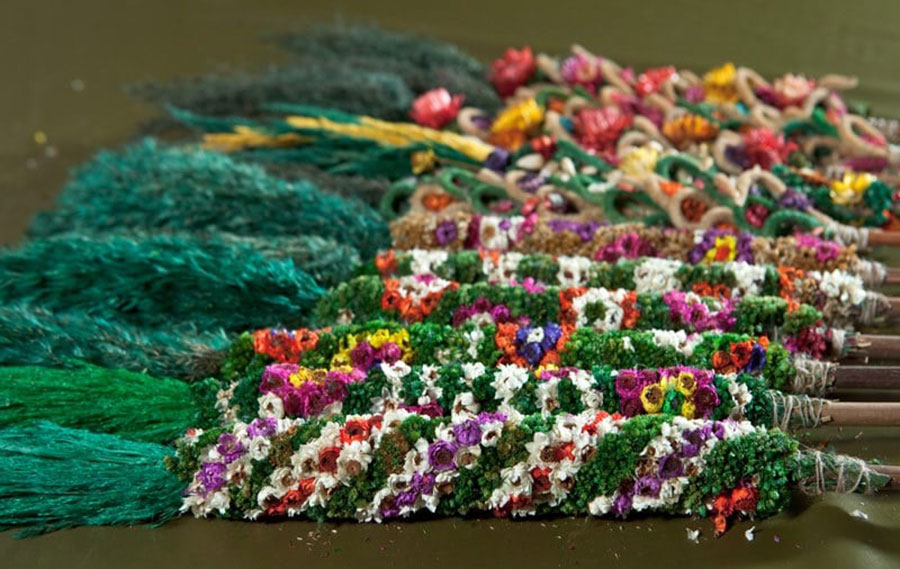 Tradycyjne palmy wielkanocne wielokolorowe wykonane z suszonych, farbowanych kwiatów, z zieloną kitą z trzciny w kolorze zielonym, ułożone jedna obok drugiej na zielonym tle