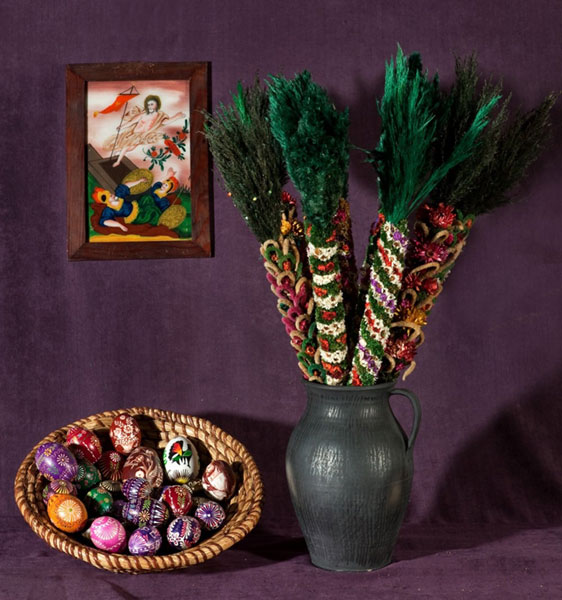 Na fioletowym tle obraz namalowany na szkle przedstawiający Zmartwychwstałego Jezusa, koszyk z jajami w wielu kolorach, ozdobione różnymi technikami, oraz ceramiczny dzban z palmami z suszonych kwiatów