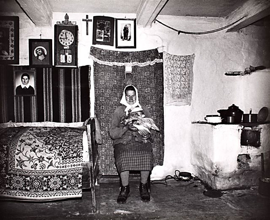 na czarno-białej fotografii w pokoju na tle wiszących kilimów pozuje starsza pani w chustce na głowie